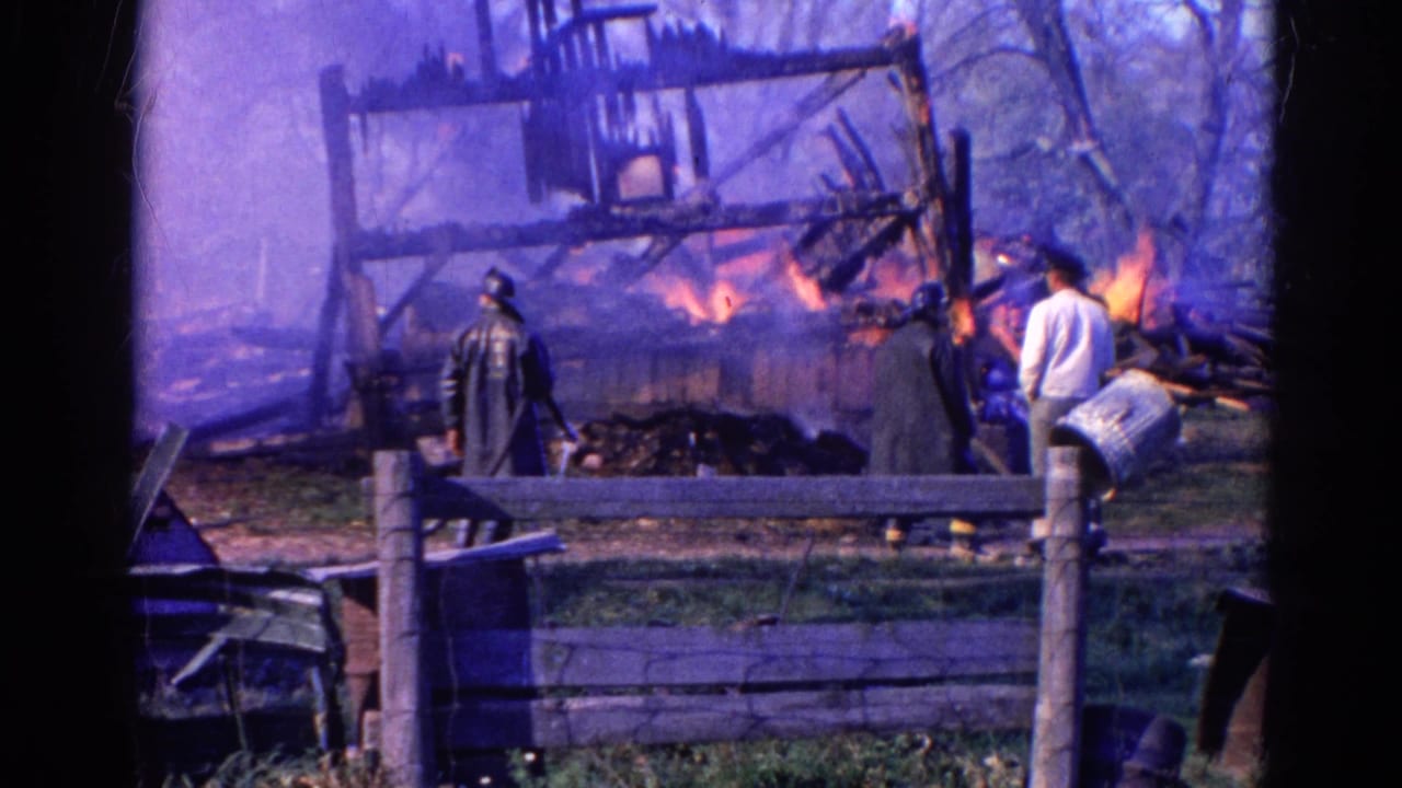 1968 Wauconda, Illinois 8mm frame showing flames, smoke, a firefighter in dark gear and bystanders watching from behind a wooden fence.
