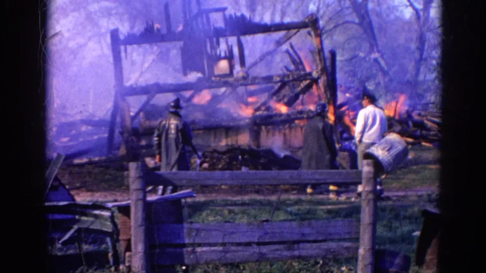 A 1968 Wauconda fire scene with a firefighter, nearby bystanders and a partially collapsed wooden structure burning behind a fence.