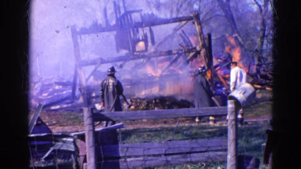 Smoke-softened 8mm frame from Wauconda, Illinois in 1968 showing a firefighter, bystanders and flames among charred wooden debris.