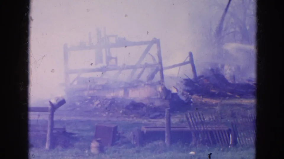 1968 Wauconda, Illinois home-movie frame showing a damaged wooden frame, scattered debris, low fencing, and a purple-blue color cast.