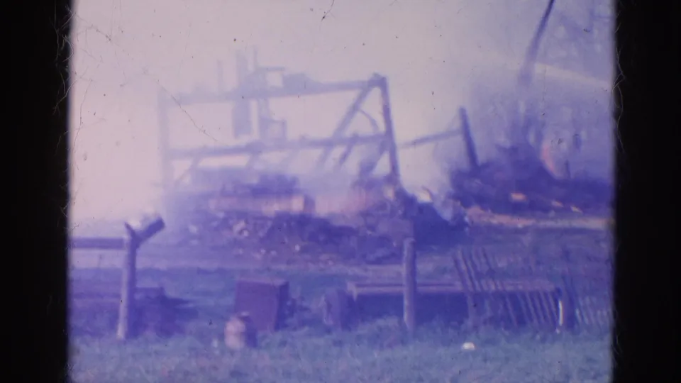 1968 Wauconda, Illinois 8mm frame of a charred structure, debris piles, fencing, grass, and bare trees in soft faded color.