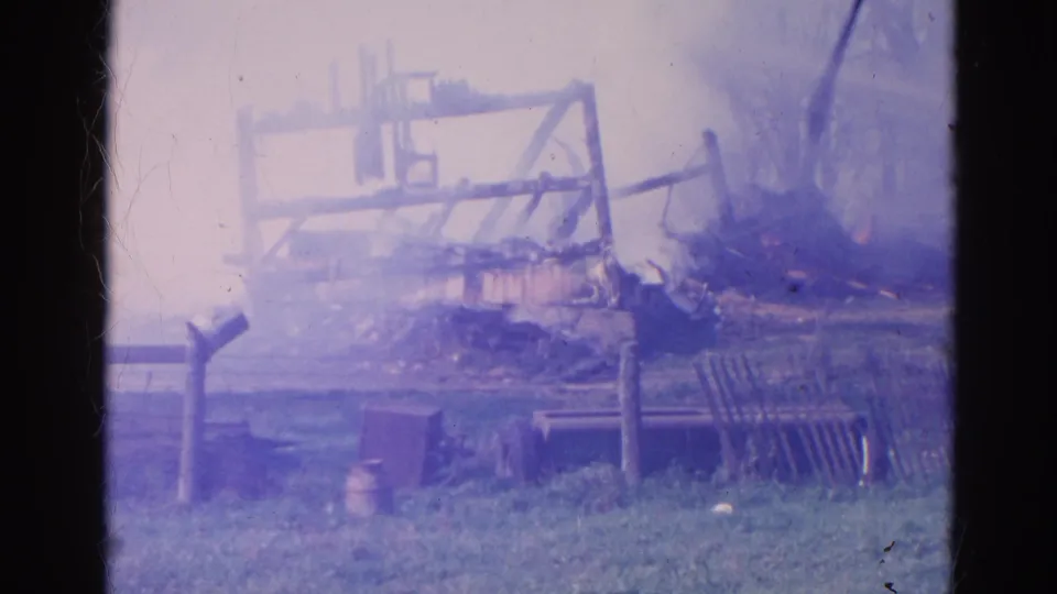 1968 Wauconda home-movie frame with smoke rising from a burned agricultural structure behind fencing and grass.