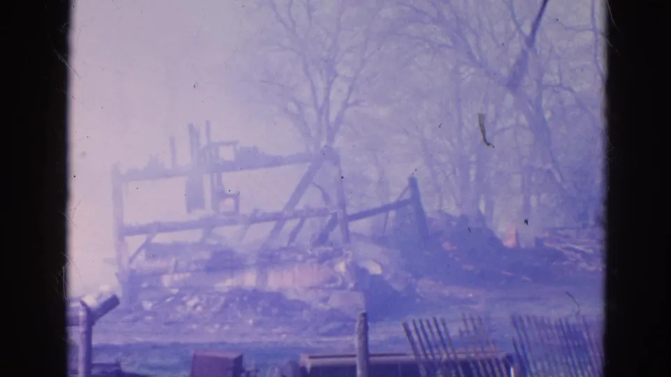 1968 Wauconda frame showing the skeletal remains of a burned rural building, smoke, trees, fencing, and foreground grass.