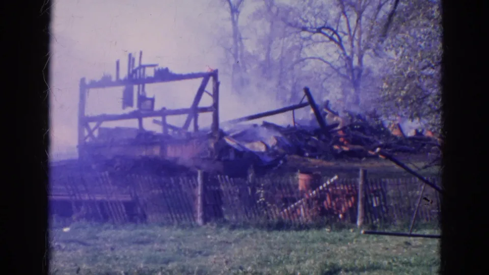 A purple-tinted 8mm frame shows the barn's charred wooden frame, scattered debris, smoke, fencing, and trees in Wauconda, Illinois.