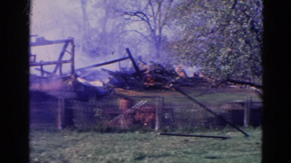 The 8mm frame shows collapsed barn debris and lingering smoke behind a fence, with open grass and leafy trees around the Wauconda fire scene.