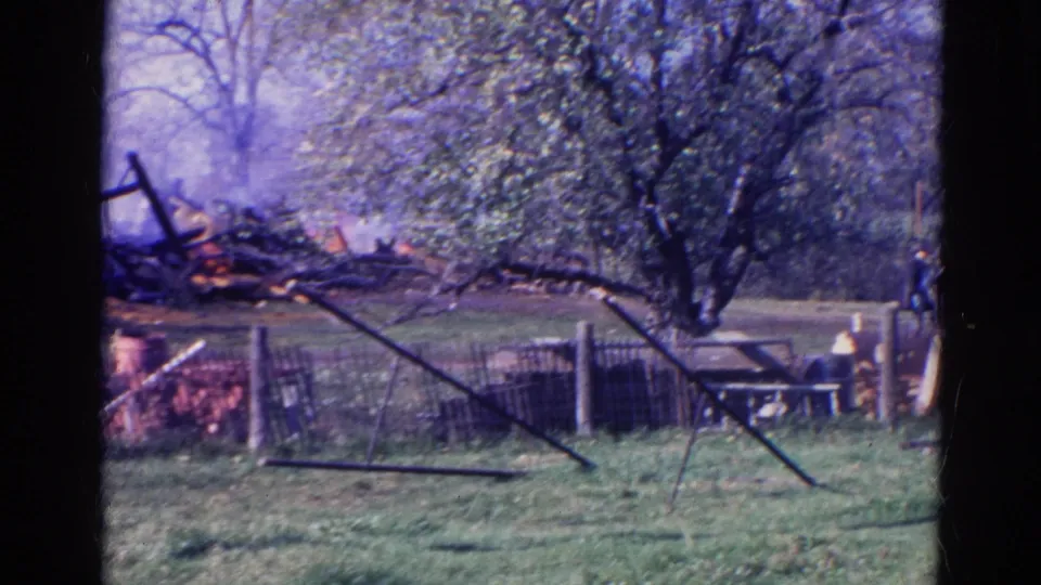 A Wauconda 1968 home-movie frame shows smoking barn wreckage, a fence, trees, and small distant bystanders near the right side.