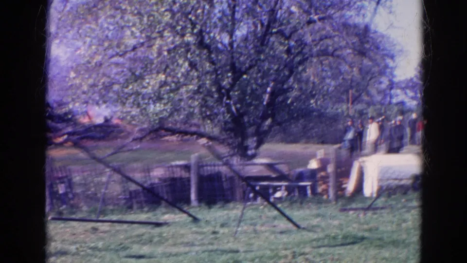 1968 Wauconda home-movie frame showing adults gathered outdoors by fencing, trees, and a light-colored yard structure.