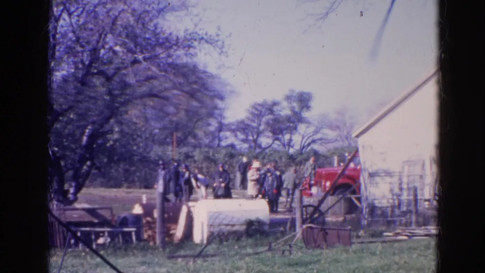 1968 Wauconda yard scene with adults near a light-colored building, fencing, trees, and red equipment.