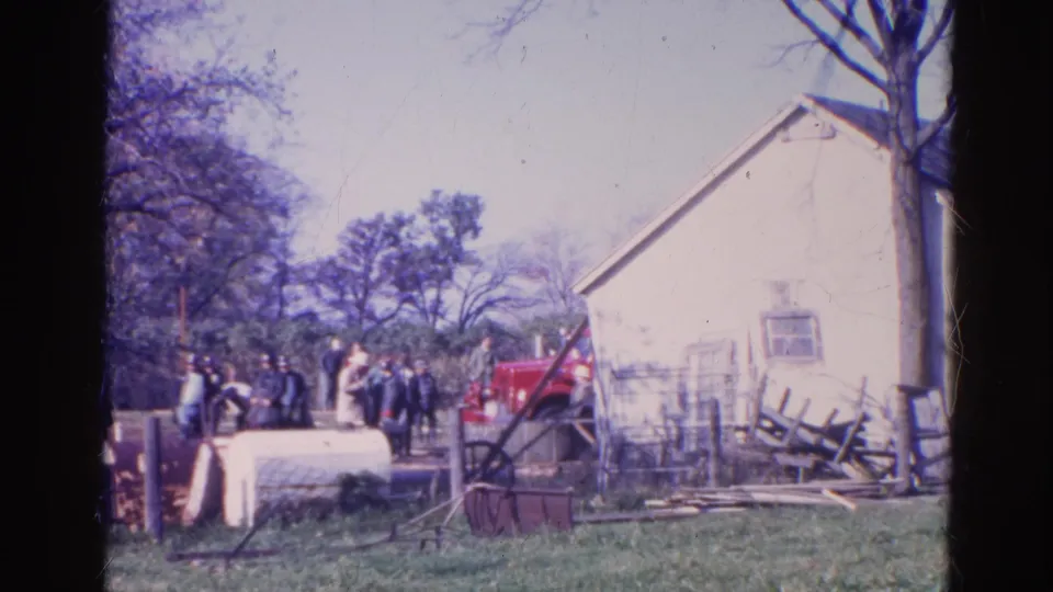 Adults in 1968 Wauconda stand in a grassy yard near a white building, red tractor-like vehicle, fencing, and trees.