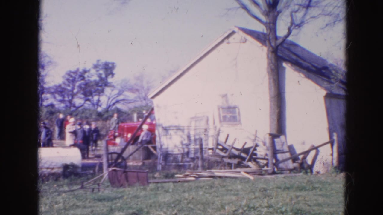 White house beside a grassy yard with onlookers, a red vehicle, and scattered fencing or debris.