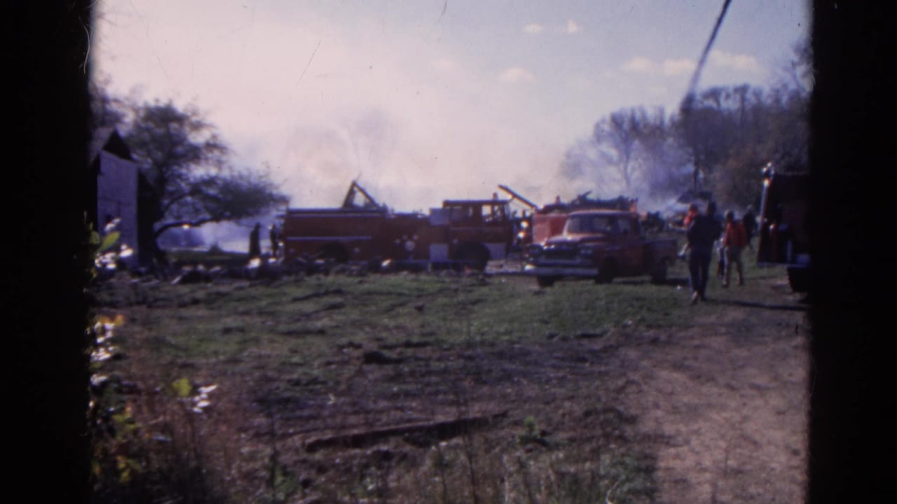 Red fire trucks and smoke in a grassy area near buildings and several people.