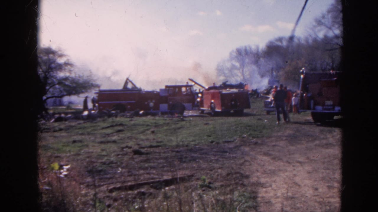 1968 Wauconda, Illinois 8mm frame showing red fire trucks, firefighters, smoke, bare trees, and a muddy field.