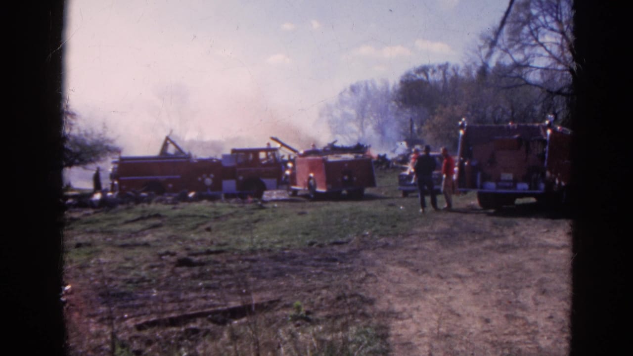 Several red fire trucks line a field while smoke rises behind them.