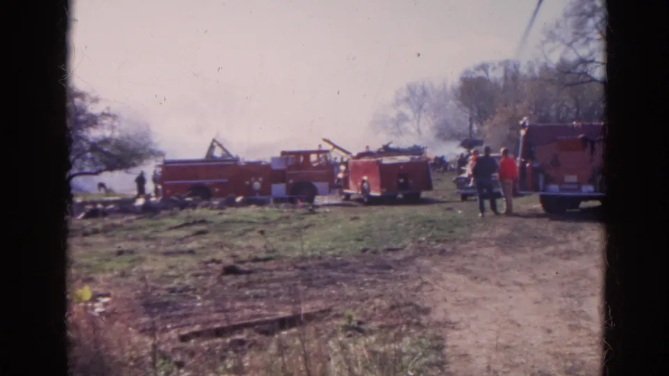 1968 Wauconda, Illinois home-movie frame showing firefighters or onlookers near red trucks and smoke in a grassy area.
