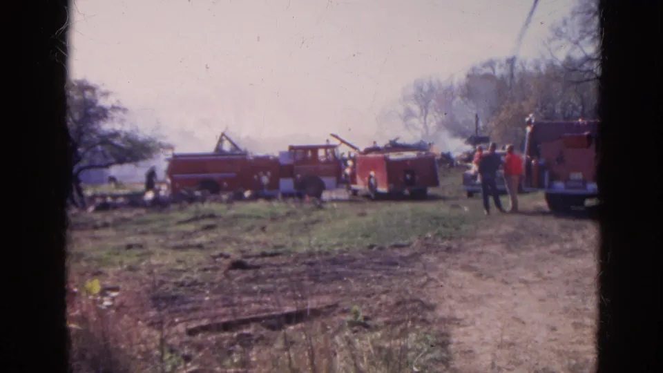 1968 Wauconda, Illinois 8mm frame showing fire trucks, distant people, drifting smoke, and a dirt access path.