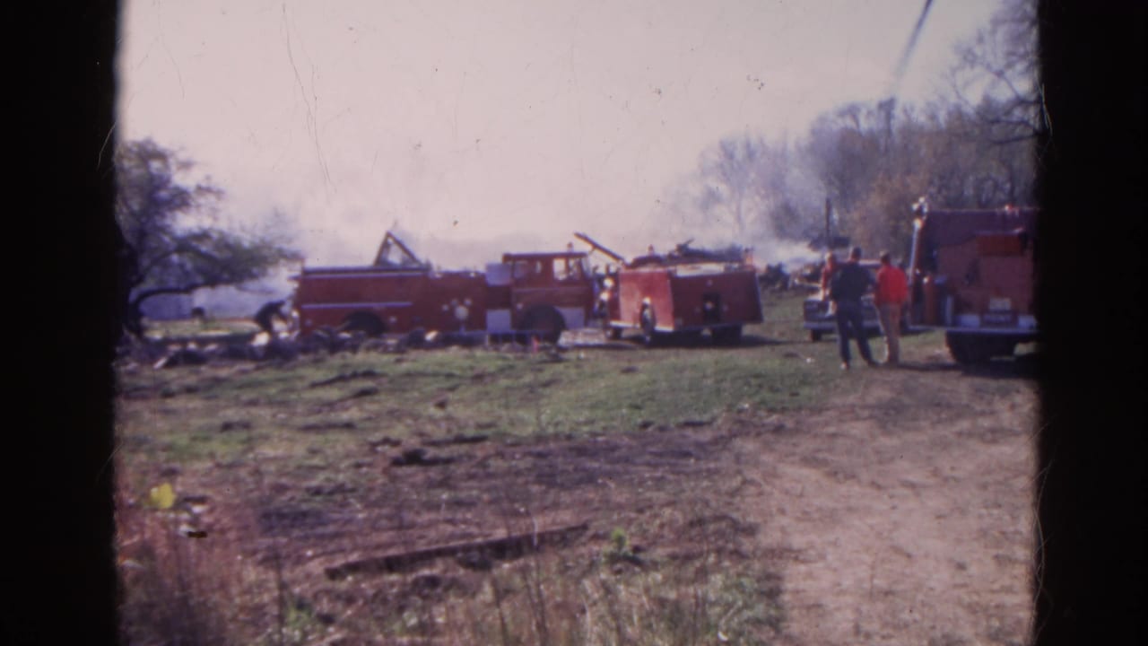 Red fire trucks parked near smoke and debris in a grassy field.