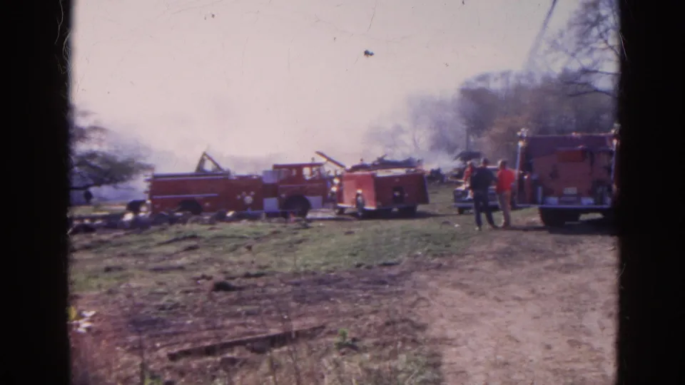 1968 Wauconda, Illinois frame with red fire trucks, distant firefighters or onlookers, smoke, and debris near trees.