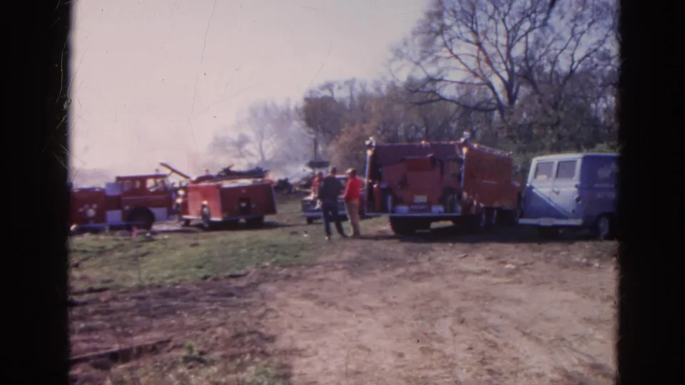 1968 Wauconda, Illinois 8mm frame showing fire trucks, two people, a blue van, smoke, and debris in a field.