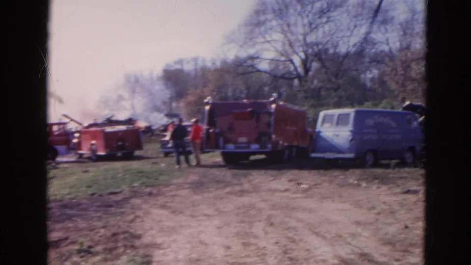 1968 Wauconda, Illinois frame with a red fire truck, blue van, people, smoke, debris, and bare trees.