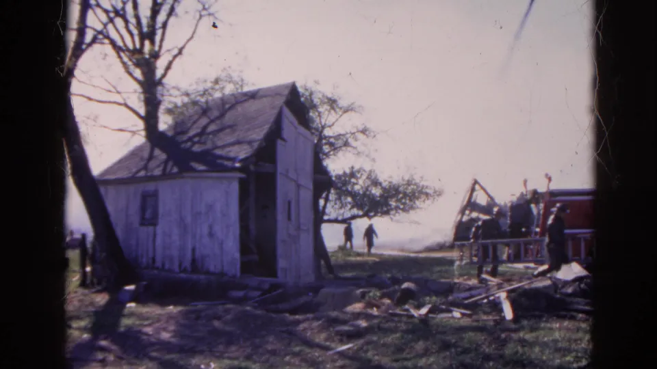 1968 Wauconda home-movie frame showing a small shed, debris, bare trees, and responders near a fire truck.