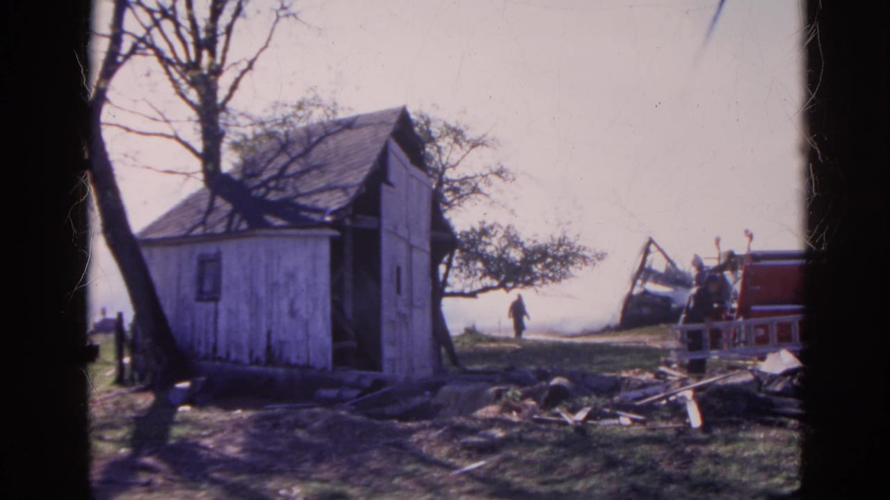 Weathered white shed with debris and distant people near a red vehicle.