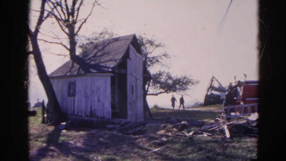 1968 Wauconda, Illinois frame with a weathered shed, two distant walkers, debris, and red equipment beside a damaged structure.