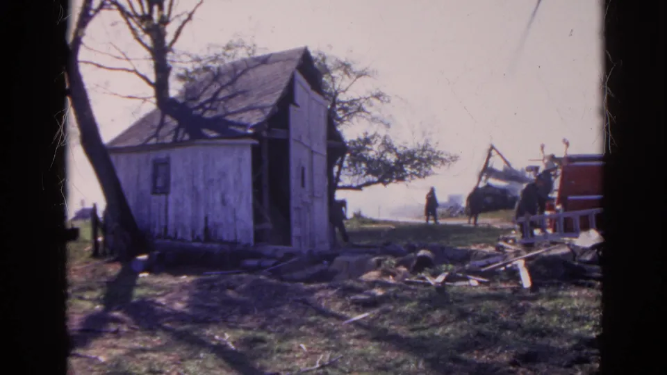 1968 Wauconda, Illinois frame of a rural shed, yard debris, bare trees, and distant people near red equipment or a vehicle.