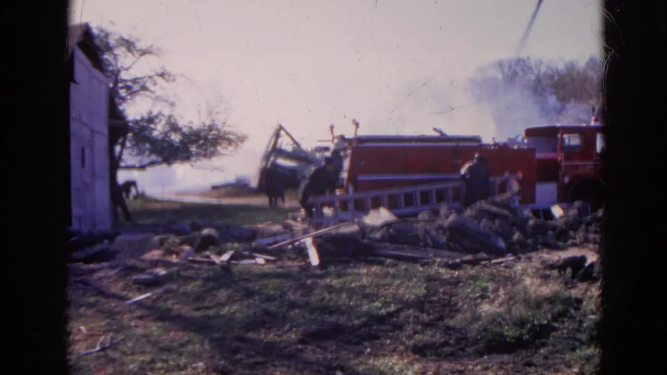 1968 Wauconda, Illinois frame with firefighters by a red fire truck, ladder, smoke, debris, and a partial shed at left.