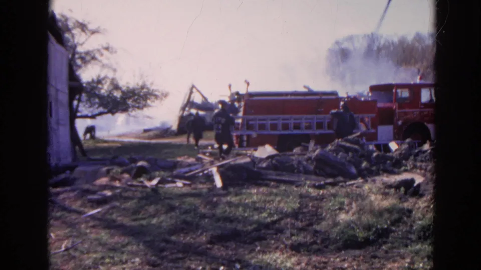 1968 Wauconda, Illinois 8mm frame showing a red fire truck, firefighters, smoke, scattered debris, and the edge of a white shed.