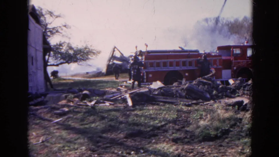 1968 Wauconda, Illinois frame showing firefighters, a red fire truck, debris, smoke, grassy ground, and a partial white shed.