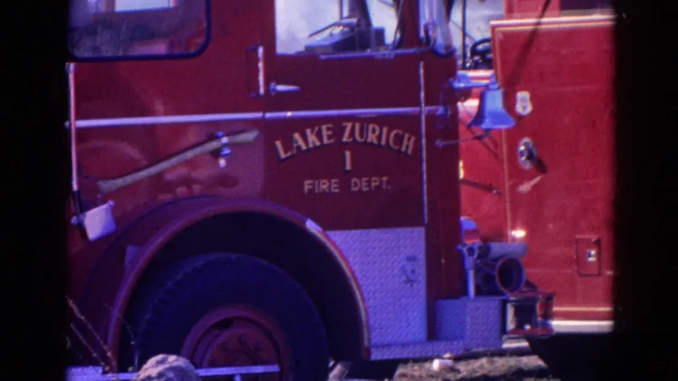 Close home-movie frame of the red Lake Zurich Fire Dept. truck side, showing lettering, wheel, hose, and equipment details.