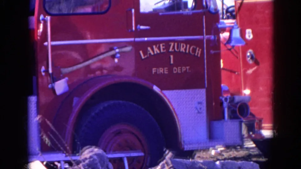 1968 home-movie frame of the Lake Zurich Fire Dept. truck, showing the side lettering, open cab area, large wheel, bell, and front hose hardware.