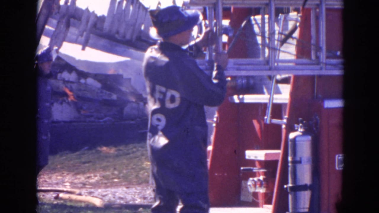 Firefighter beside a red fire truck and ladder with damaged structure behind.