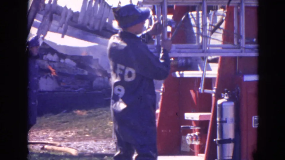 1968 Wauconda home-movie frame showing a firefighter handling a ladder near a red fire truck as debris and flames appear behind.