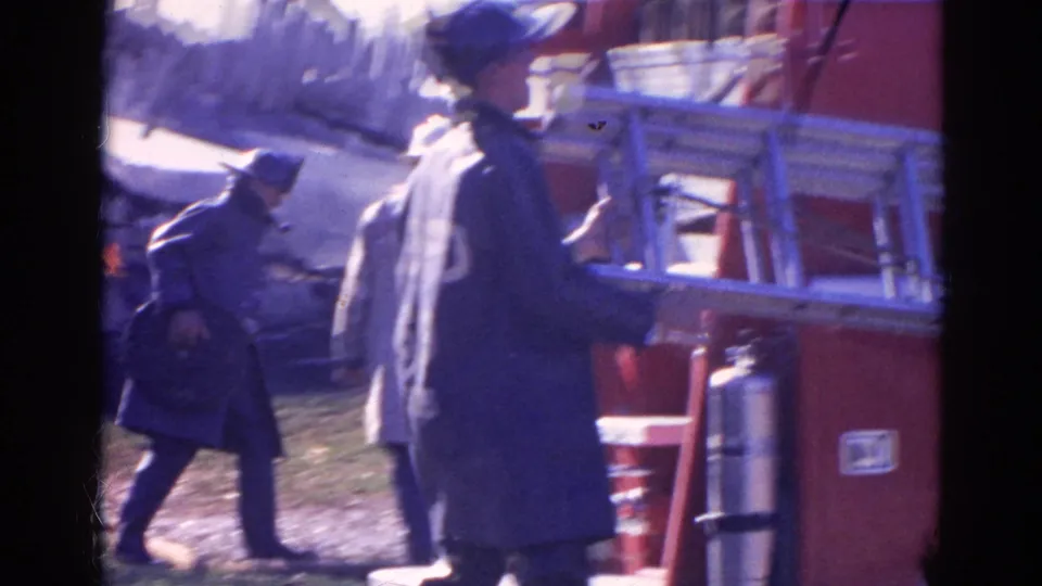 Two firefighters at the Wauconda fire scene move near a red truck and ladder, with damaged material visible behind them.