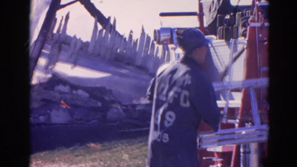 A firefighter with visible coat lettering stands by a ladder near the red fire truck, with smoldering debris in the background.