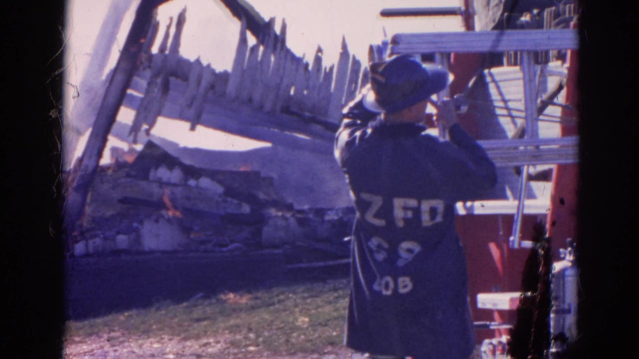 In 1968 Wauconda, Illinois footage, a helmeted firefighter stands by red apparatus facing a burning damaged structure and scattered debris.
