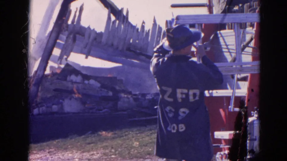 In 1968 Wauconda, Illinois footage, a helmeted firefighter stands by red apparatus facing a burning damaged structure and scattered debris.