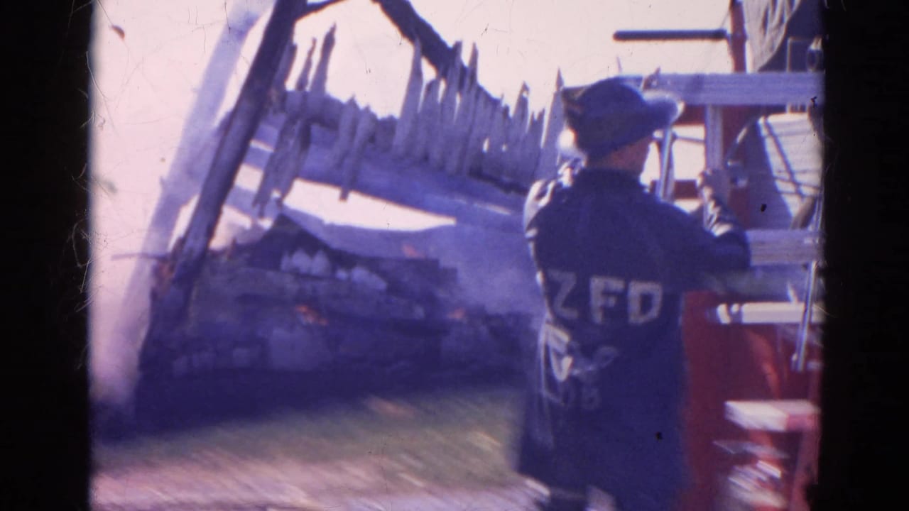 A firefighter seen from behind stands near red fire equipment as smoke, flames, and damaged material appear in the background.