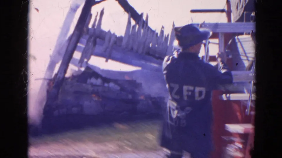 A firefighter seen from behind stands near red fire equipment as smoke, flames, and damaged material appear in the background.