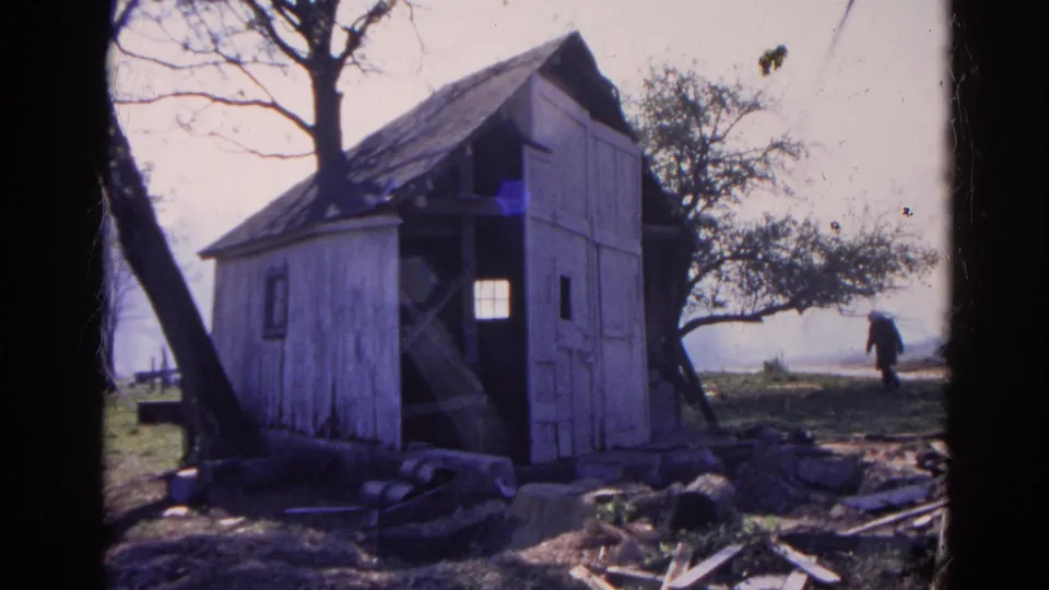 The rural shed remains in view while a distant person walks near trees and open ground, with debris scattered in the foreground.