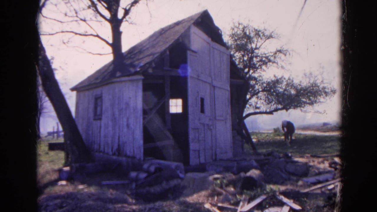 1968 Wauconda, Illinois home-movie frame of a weathered white outbuilding, leafless trees, debris, and a man bending near the horizon.