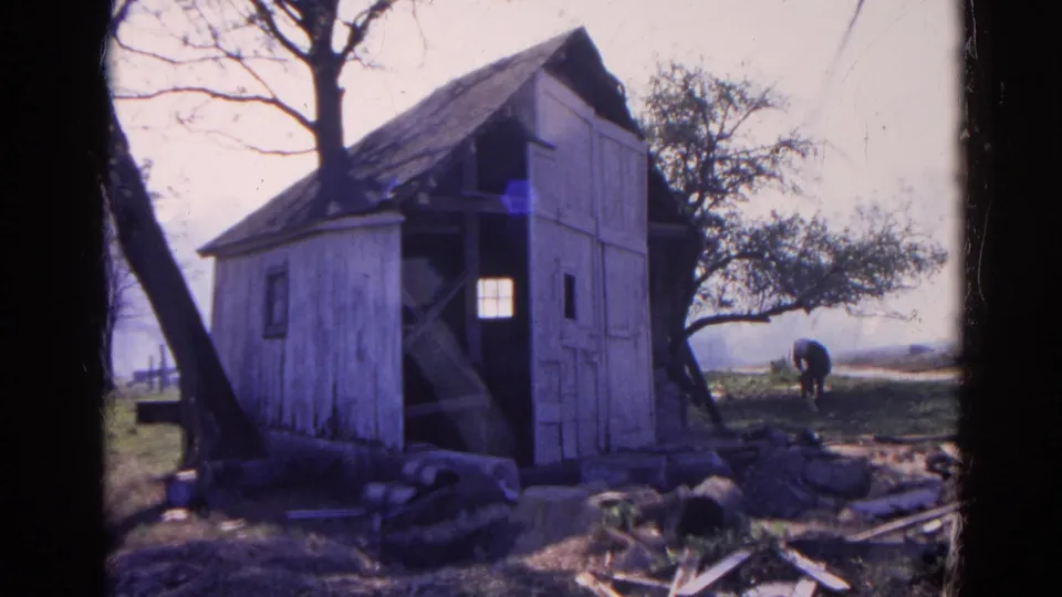 1968 Wauconda, Illinois home-movie frame of a weathered white outbuilding, leafless trees, debris, and a man bending near the horizon.