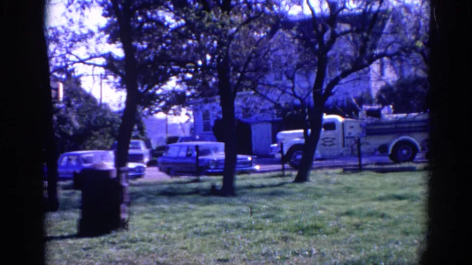 Final 1968 Wauconda park frame with blue-cast 8mm texture, trees in the foreground, parked cars, and a white utility truck behind them.