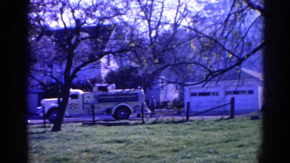 The 1968 home-movie frame shows a white fire truck beside a residential garage, with trees, grass, and a small figure nearby.
