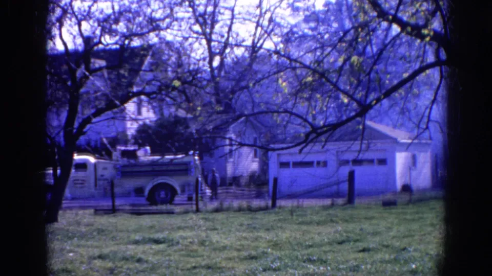 A late-1960s Wauconda frame with a white fire truck beside a garage, tree branches overhead, grass in front, and residential buildings behind.