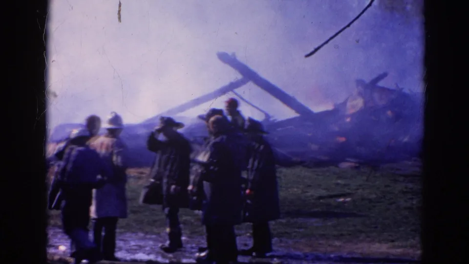Firefighters in coats and helmets approach smoking wreckage in the 1968 Wauconda fire scene.