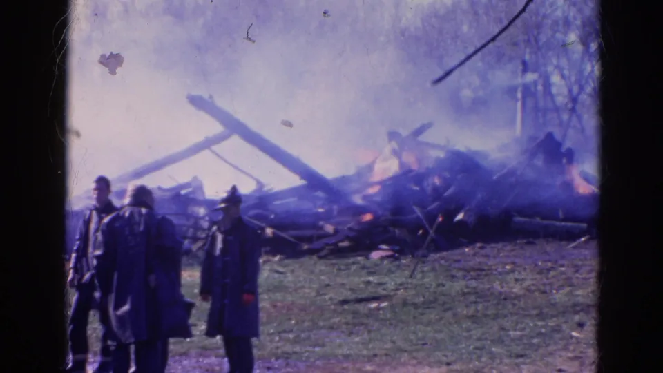 Two firefighters in period coats stand near smoke, charred material, and visible flames in Wauconda, Illinois.