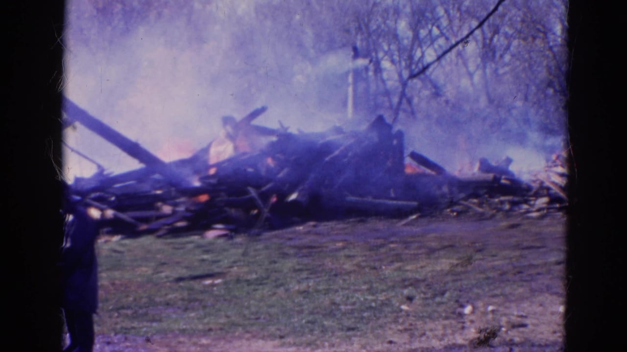 Debris pile burning in a grassy field with smoke and a person standing at left.