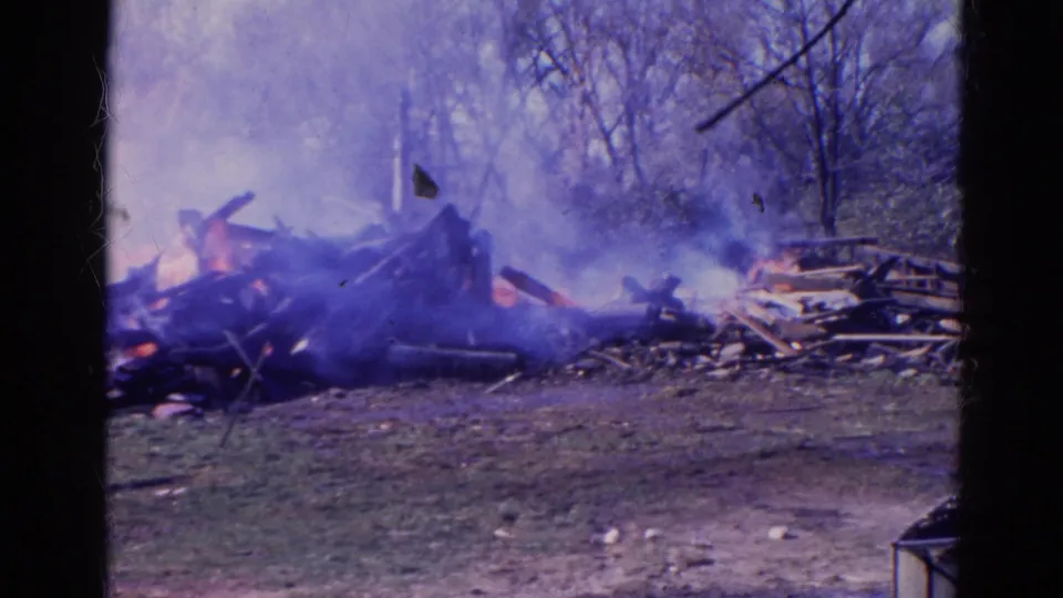 1968 Wauconda, Illinois frame of a debris fire sending smoke across a field, with leafless trees and wooden wreckage visible.